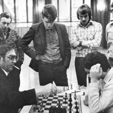 Fólk stúgvast kring borðið at forvitnast. Føroyar og Ísland telva landsdyst í auluni í Hoydølum í 1976. Telvarin vinstrumegin er Petur Eli Mikkelsen úr Gøtu Talvfelag, ið sat á 2. borði (Mynd: Talvsamband Føroya)